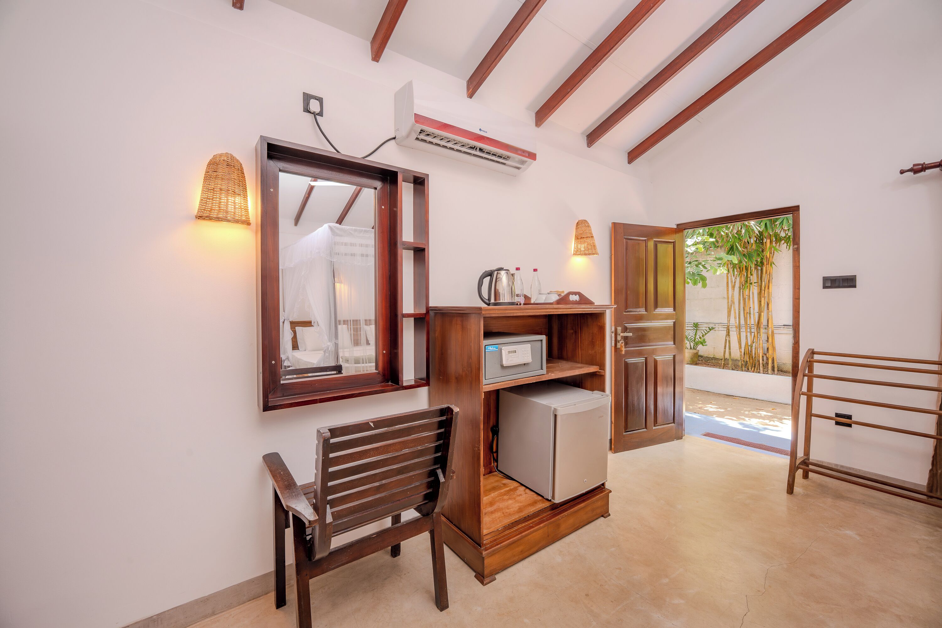 A room with a wooden-framed mirror, a small refrigerator, a wooden chair, and a shelf with a kettle and bottles.