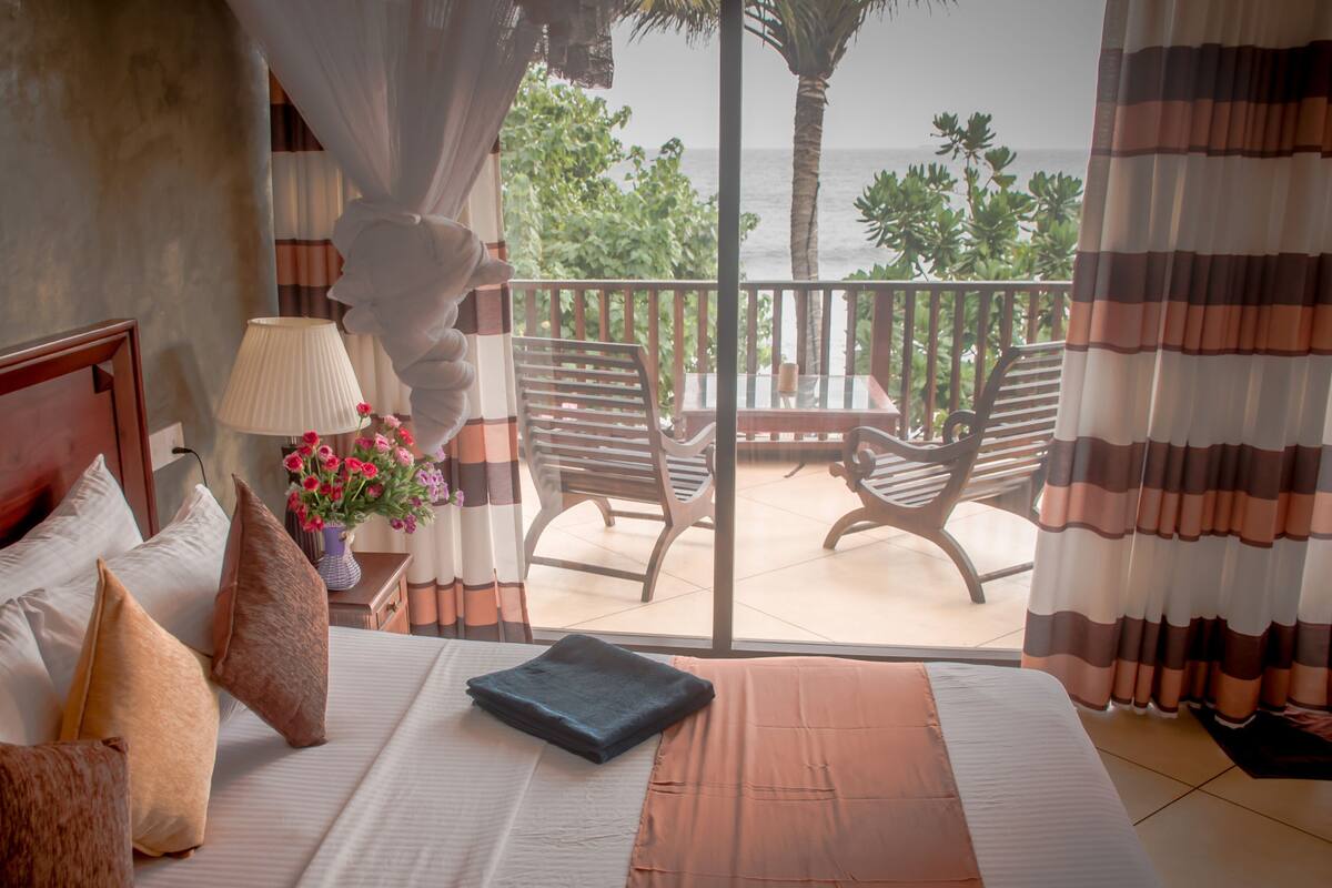 A hotel room with a bed, a nightstand with a lamp and flowers, a view of the ocean, and a balcony with chairs.