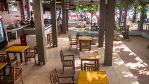 A beachside restaurant with outdoor seating, wooden tables and chairs, and red umbrellas.