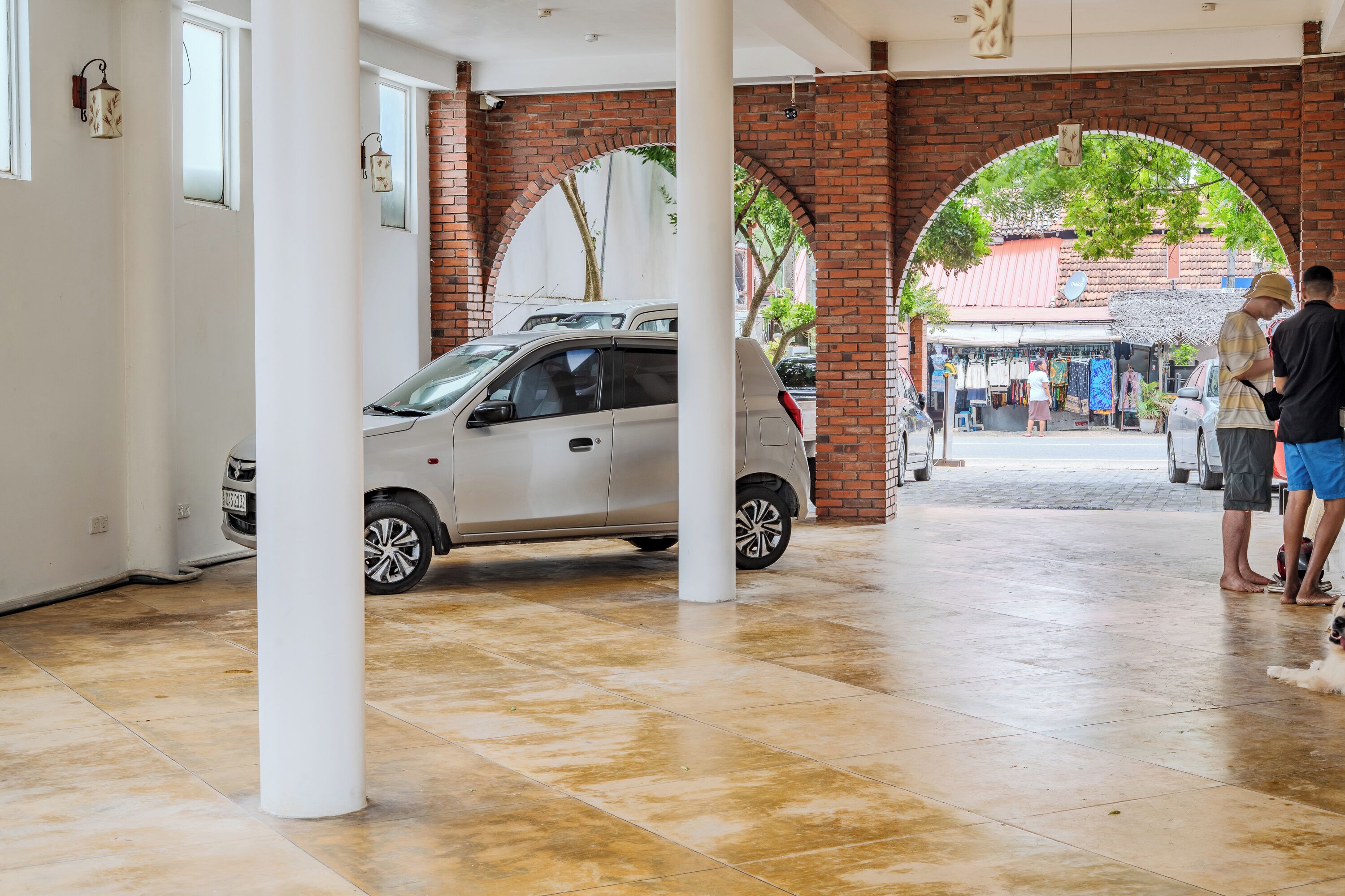 A covered parking area with a white car, brick arches, and a view of a street with shops and people.