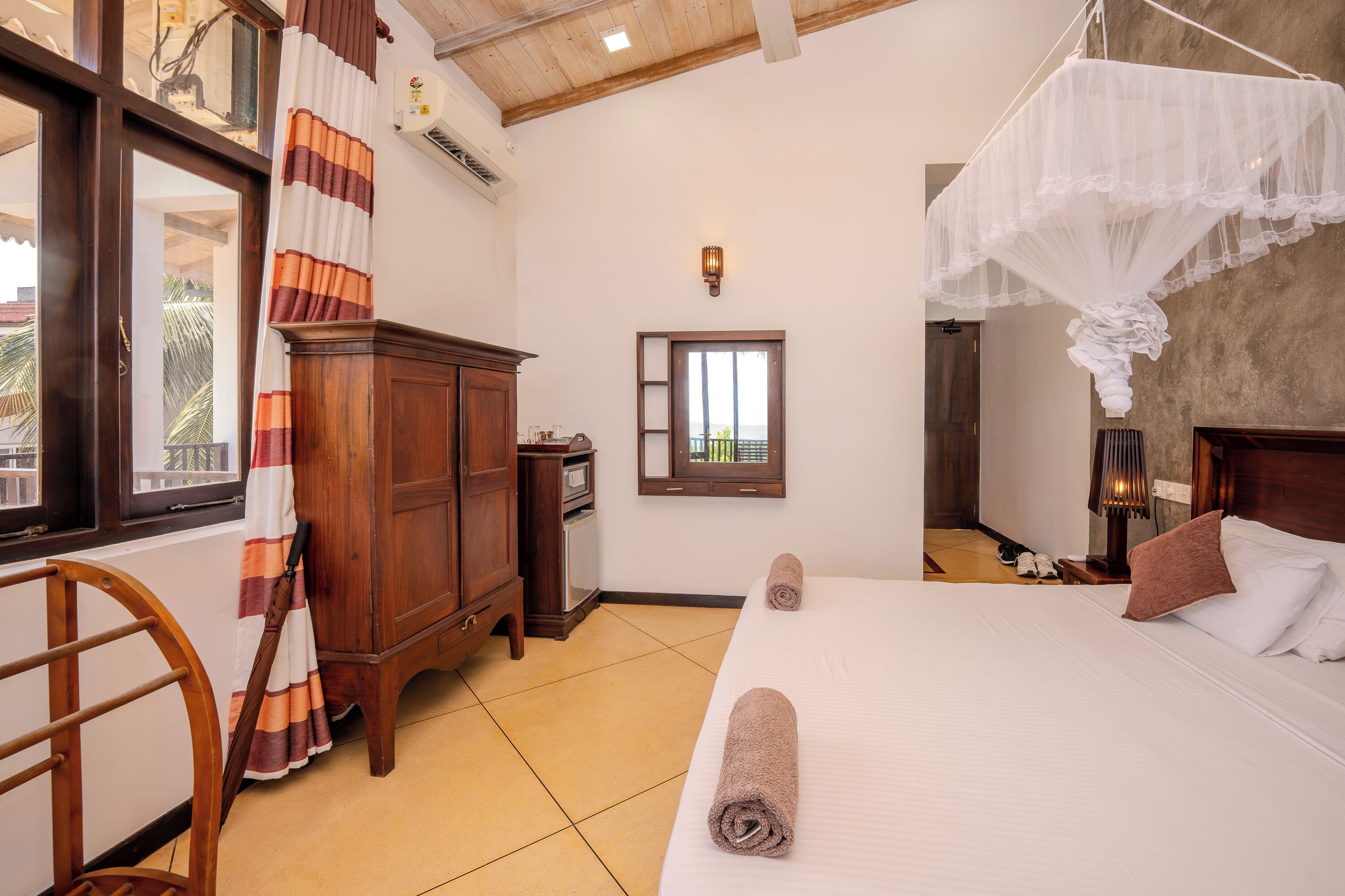 A hotel room with a bed, a wooden wardrobe, a window with curtains, and a nightstand with a lamp.