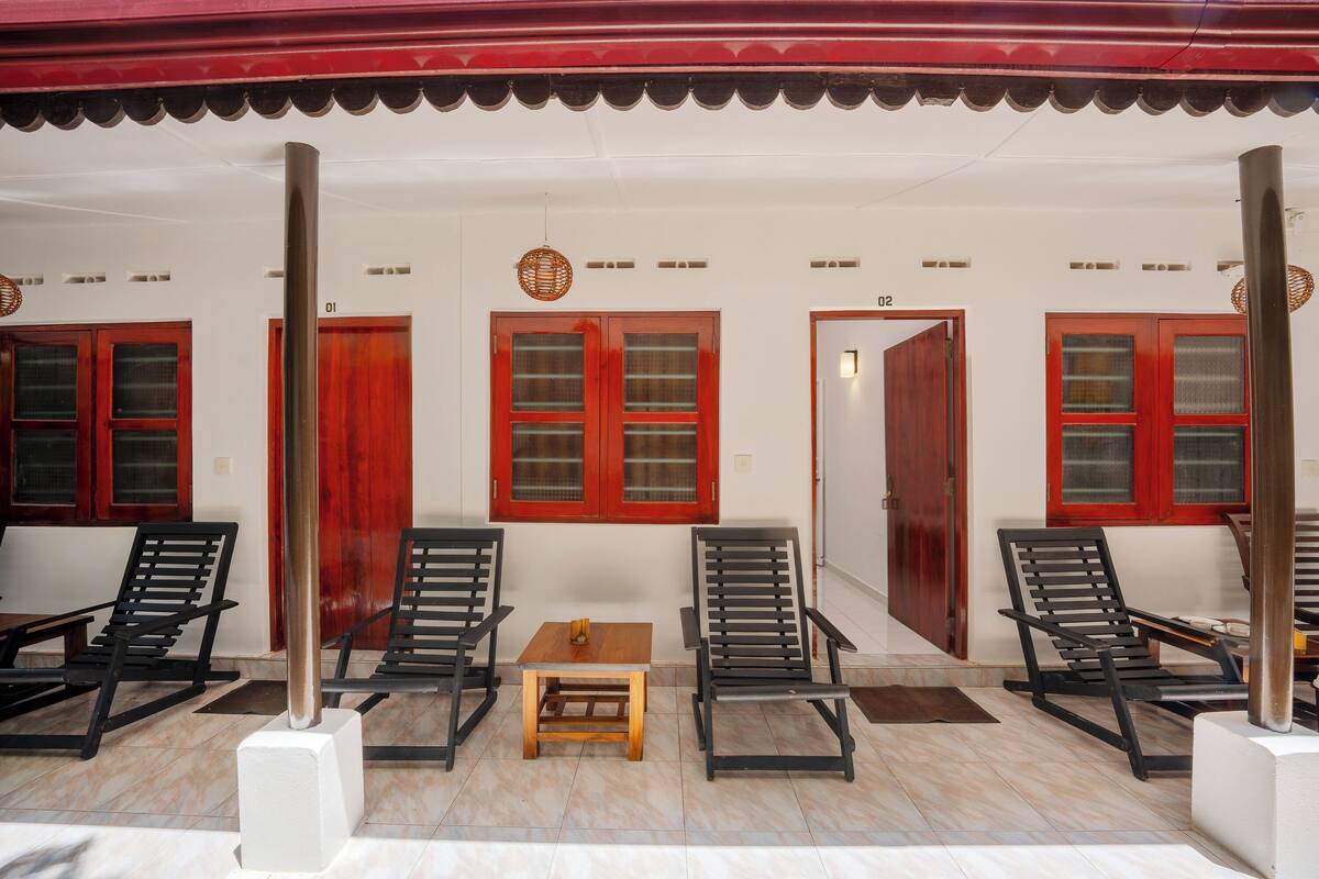 A covered outdoor seating area with wooden tables and chairs, red-framed windows, and a tiled floor.