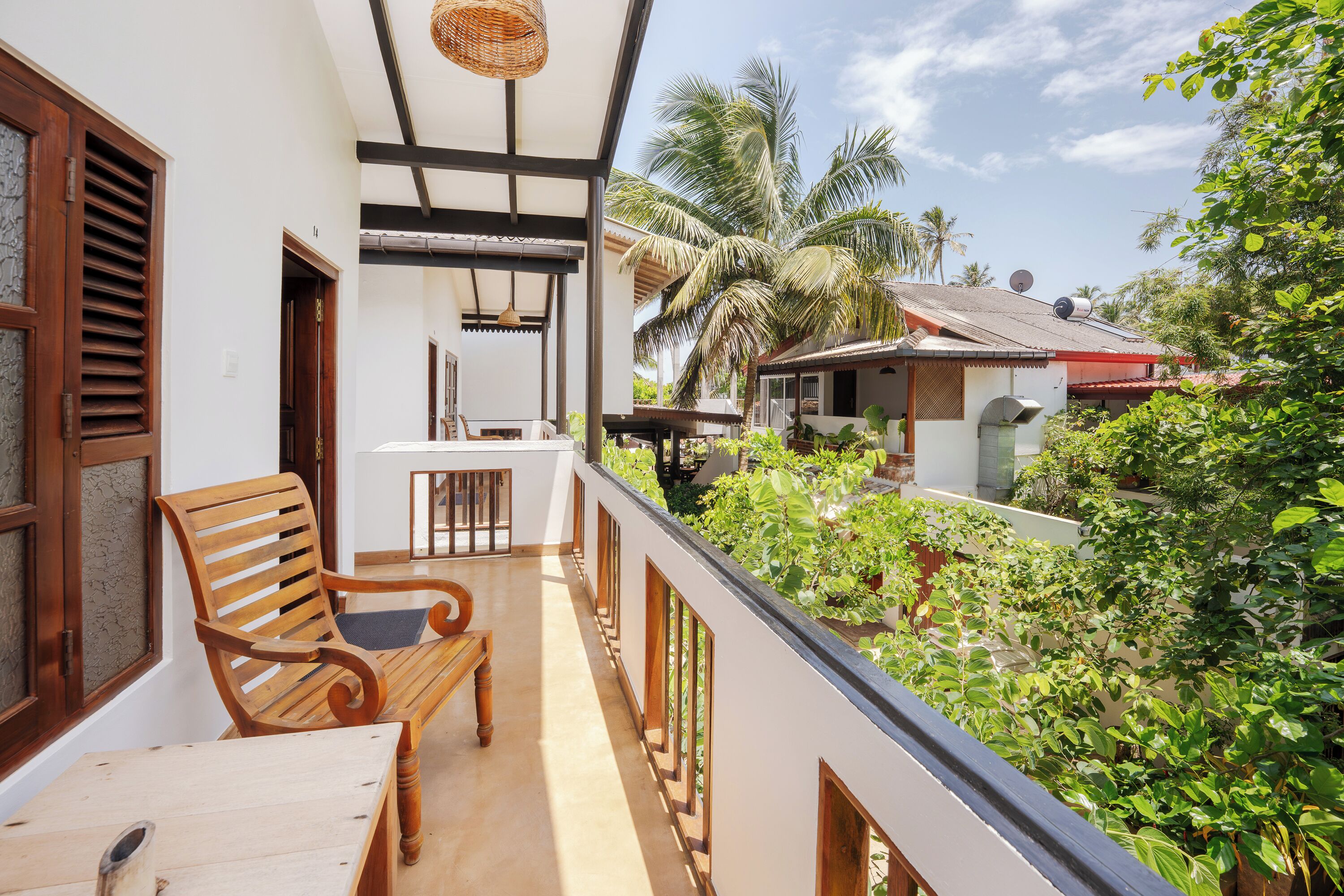 A balcony with a wooden bench and table, overlooking a lush garden and neighboring houses.