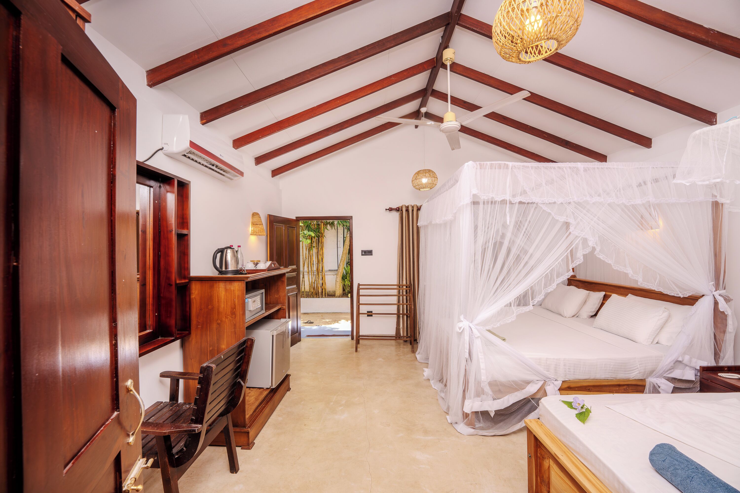 A room with a bed under a canopy, a wooden bench, a desk with a chair, and a window with curtains.