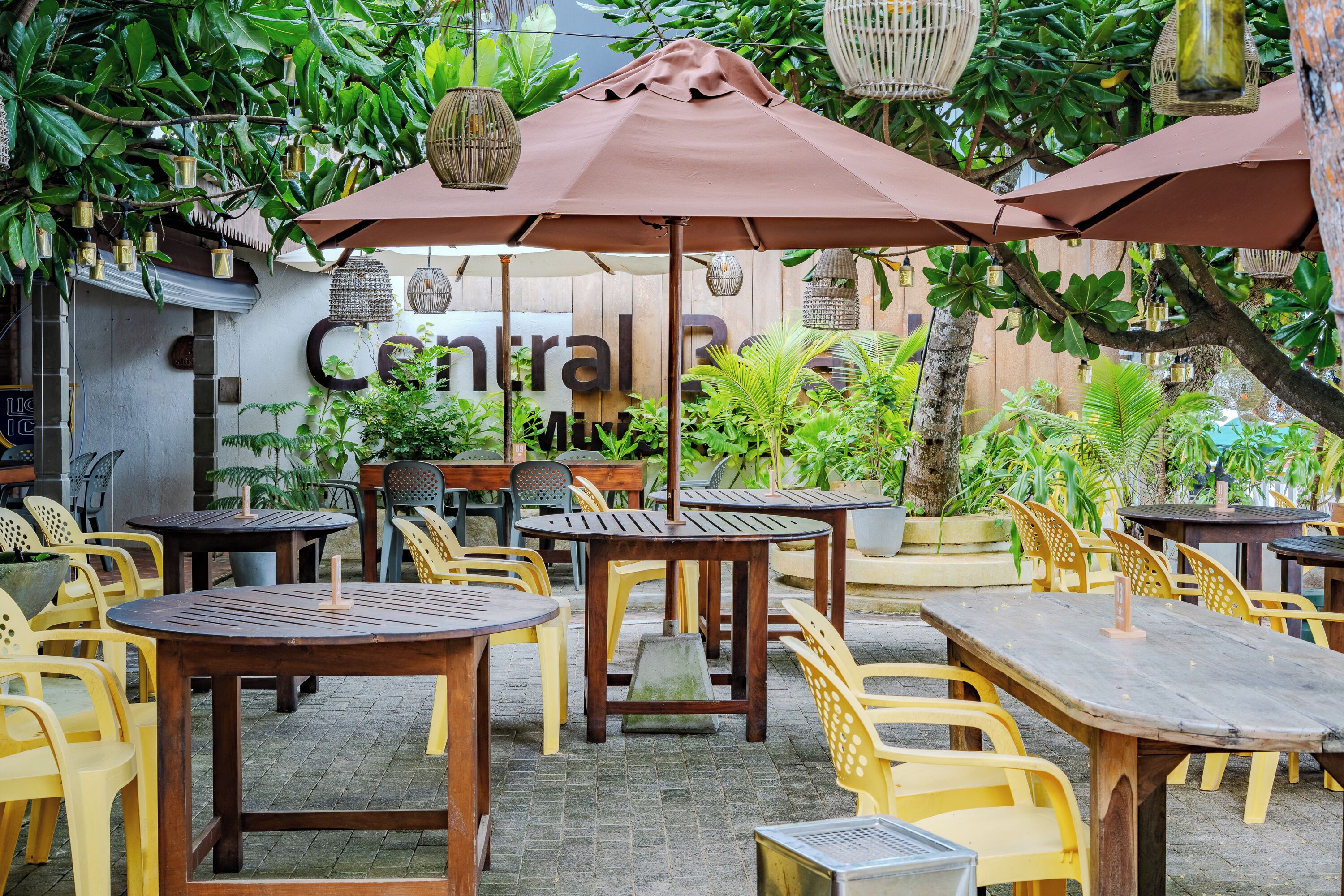 An outdoor seating area with wooden tables and yellow chairs, surrounded by greenery and hanging lanterns.