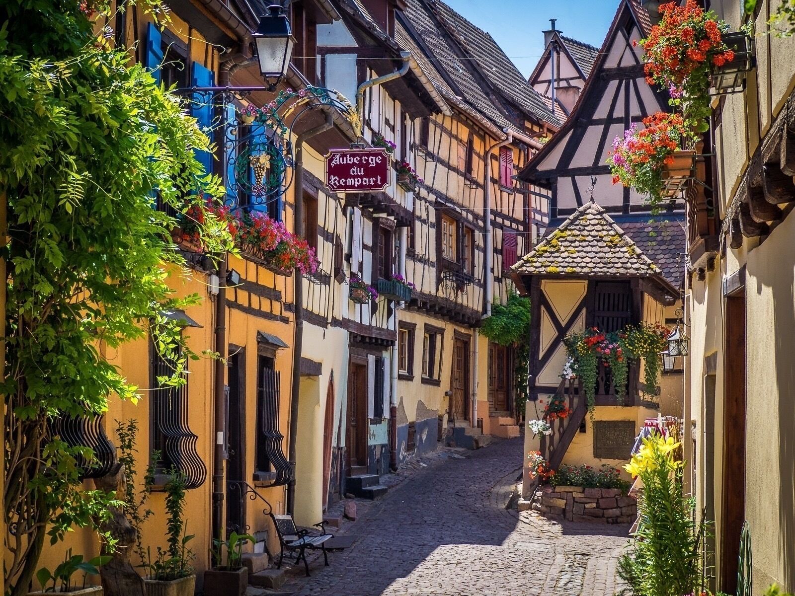 Eine enge Kopfsteinpflasterstraße gesäumt von traditionellen Fachwerkhäusern, mit einem Schild, auf dem 'Auberge du Rempart' steht, und Kübeln mit Blumen.