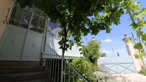 Un escalier menant à une porte blanche surmontée d’un portail en métal, le tout entouré de verdure et sous un ciel dégagé.