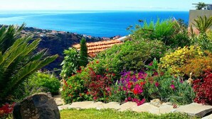 A coastal landscape with a view of the sea, a garden with colorful flowers, and a building with a red-tiled roof. - Amazing views, large terrace and garden. (Madère)
