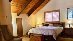 Ein Schlafzimmer mit Holzbalkendecke, einem Bett mit weißer Bettdecke, einer hölzernen Kommode, einem Stuhl und einem Fenster mit Jalousien.