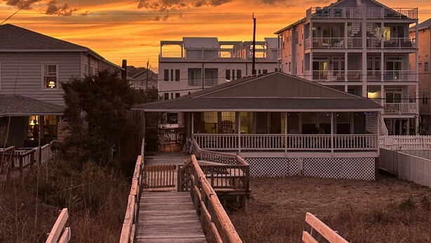 Una pasarela de madera que conduce a un edificio frente a la playa, con balcón y zona cubierta.