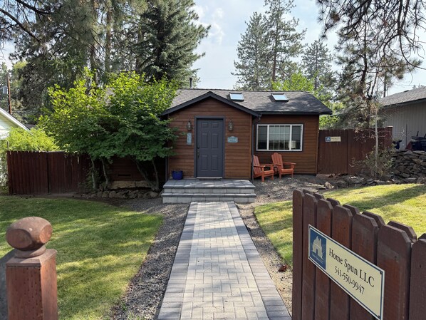 A wooden house with a paved driveway, a front door, and a small patio with chairs.