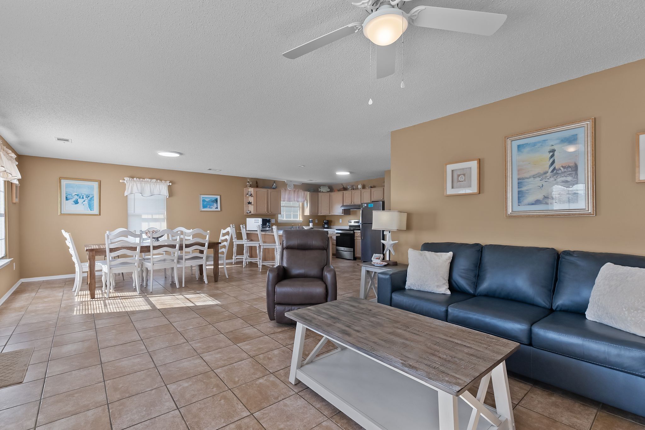 A spacious living room with a blue leather sofa, a dining area with white chairs, a brown leather chair, and a ceiling fan.