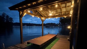 A wooden deck with a bench, a pergola with string lights, and a view of a calm lake at dusk. - Private Lakeside, private Dock, Outddor Kitchen Fire Pit & More (Chappells)