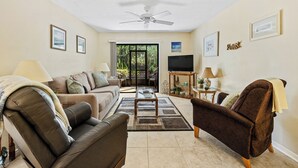 Una sala de estar con ventilador de techo, un sofá, un sillón marrón, una mesa de centro y un televisor sobre un mueble.