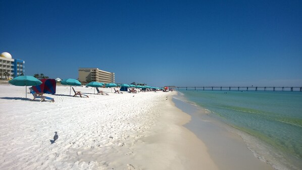 A beach with white sand, blue umbrellas, and a pier in the background.