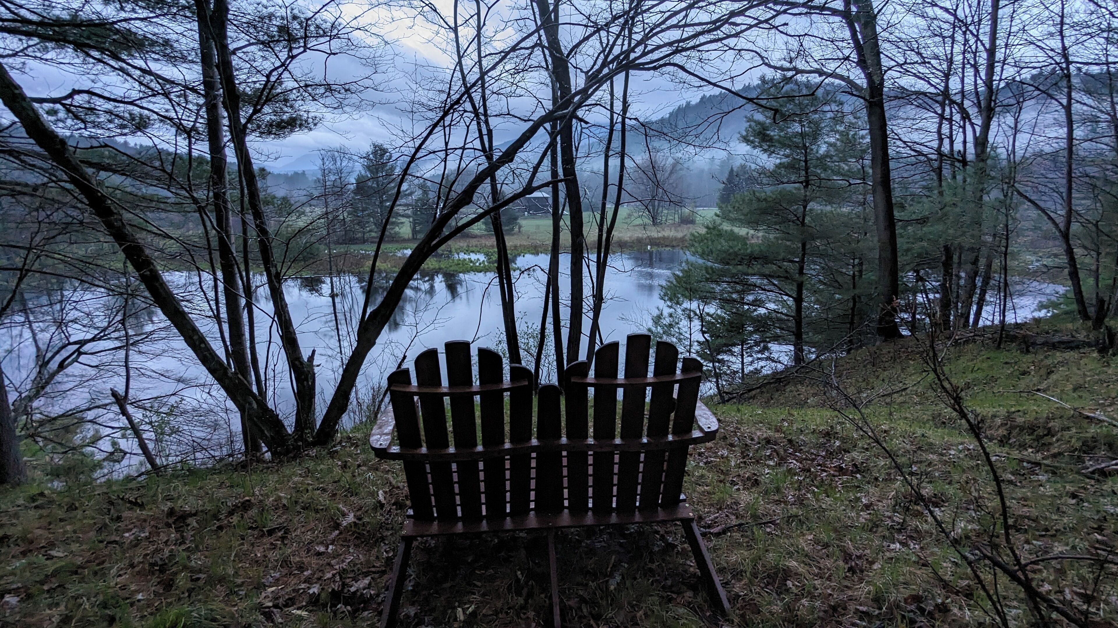 A wooden Adirondack chair facing a lake surrounded by trees.