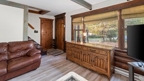 A living room with a brown leather couch, wooden cabinet, and a large window with a view of trees. - Secluded Family Getaway Cabin near Whiteface (Upper Jay)
