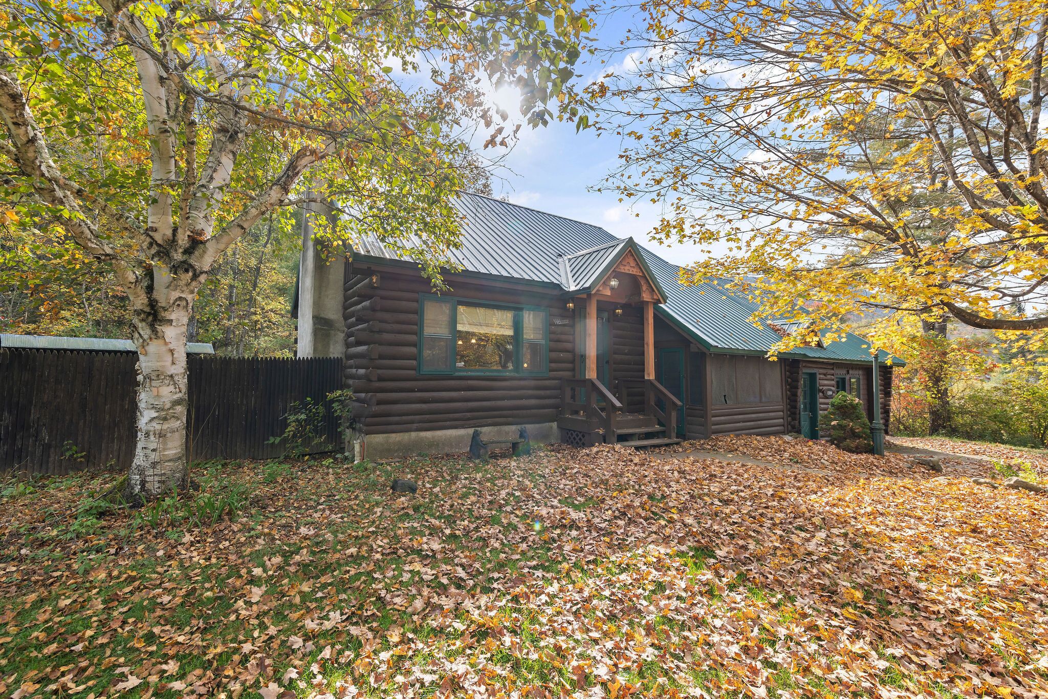 A rustic log cabin with a metal roof, surrounded by autumn leaves and trees.
