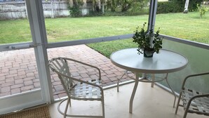 A patio area with a round table and two wicker chairs, a potted plant on the table, and a view of a grassy yard through a glass door. - Coastal Cottage near beaches, dining & shopping! (Naples)
