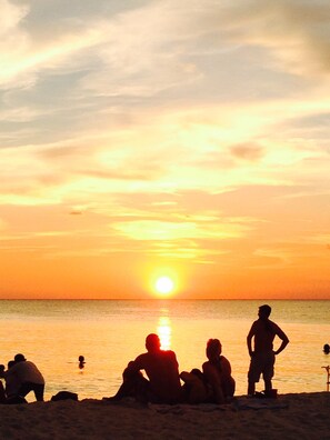 Ein Sonnenuntergang über einem Strand, Menschen sitzen und stehen im Sand.