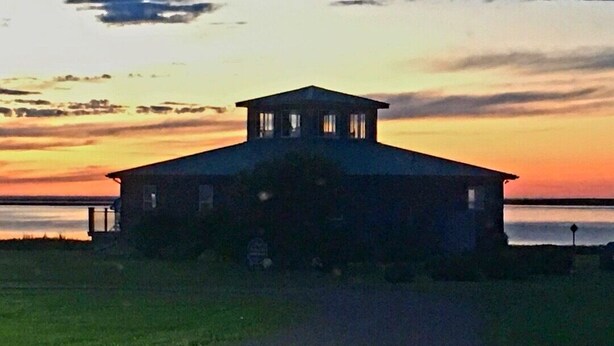 A large, single-story building with a central roof and a flat roofline, situated on a grassy area with a body of water in the background. - Ocean Edge Beach House (Malpeque)