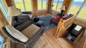 L’intérieur d’un chalet accueillant est aménagé avec un canapé en osier, une petite table et une lampe. De grandes fenêtres offrent une vue sur un paysage verdoyant et sur une étendue d’eau.