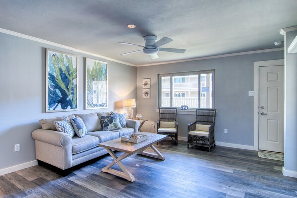 A living room with a sofa, coffee table, and chairs. There is a ceiling fan, a window, and a door.