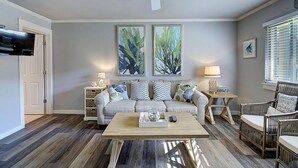 Un salon avec un canapé gris, une table basse et deux tables d’appoint avec des lampes. Il y a deux tableaux accrochés au mur et un ventilateur de plafond.