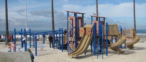A playground with various play structures, including slides and climbing areas, situated on a sandy area with a view of the sea in the background.