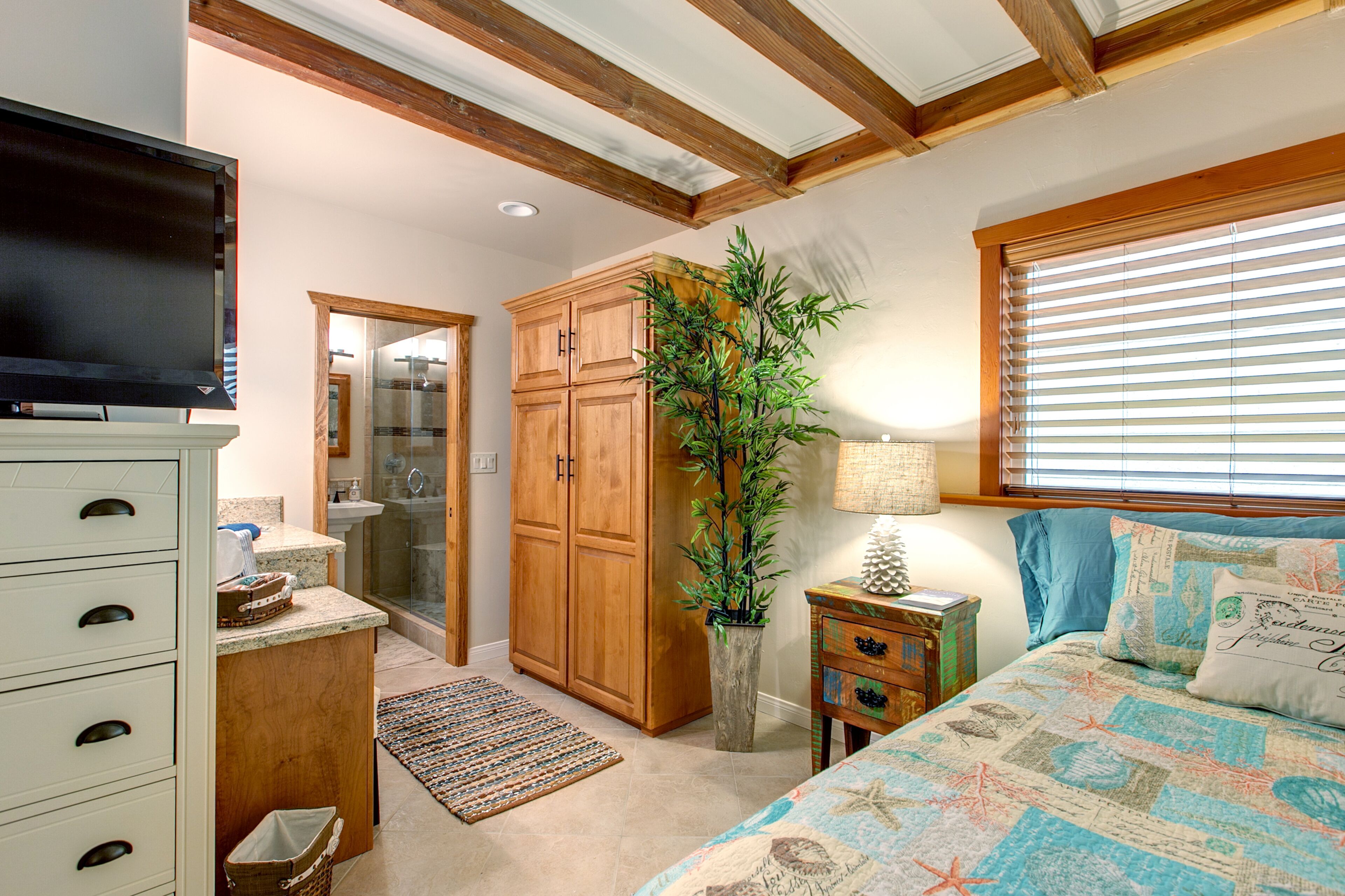 A bedroom with a bed, a TV, wooden cabinets, a plant, and a bathroom visible through an open door.