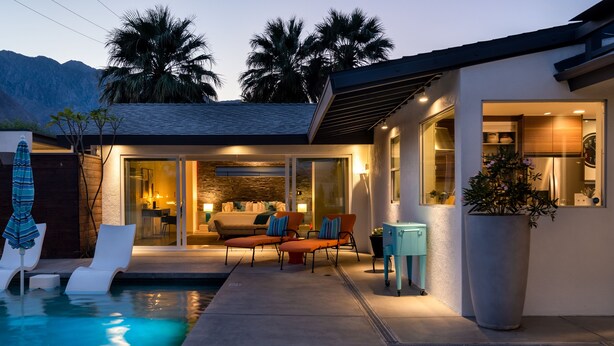 Una casa moderna con piscina, mobiliario exterior y una cocina visible a través de puertas de vidrio.