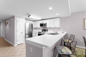 A modern kitchen with white cabinetry, stainless steel appliances, a central island, and a dining area with two chairs. - Amalfi Coast 213B - Community Pool, Fitness Facility, Gulf Views, Miramar Beach! (Miramar Beach)