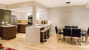 A modern kitchen with white cabinets, a central island, and a dining area with a glass table and black chairs. - Private Hot Tub & AC | Pool | Free Parking | Steps to Village, Lifts & Bike Park (Whistler)