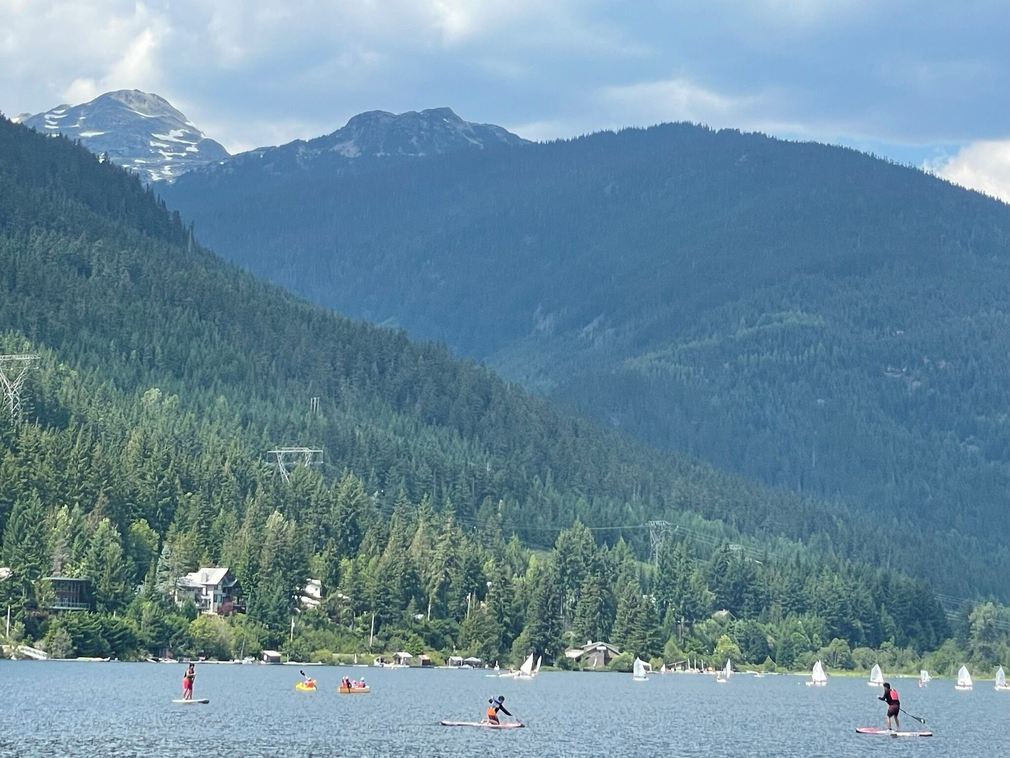 A lake with paddleboards and sailboats, surrounded by forested hills and houses.