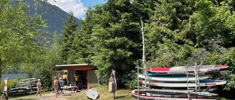 A lakeside setting with a small wooden structure, kayaks, and a paddleboard.