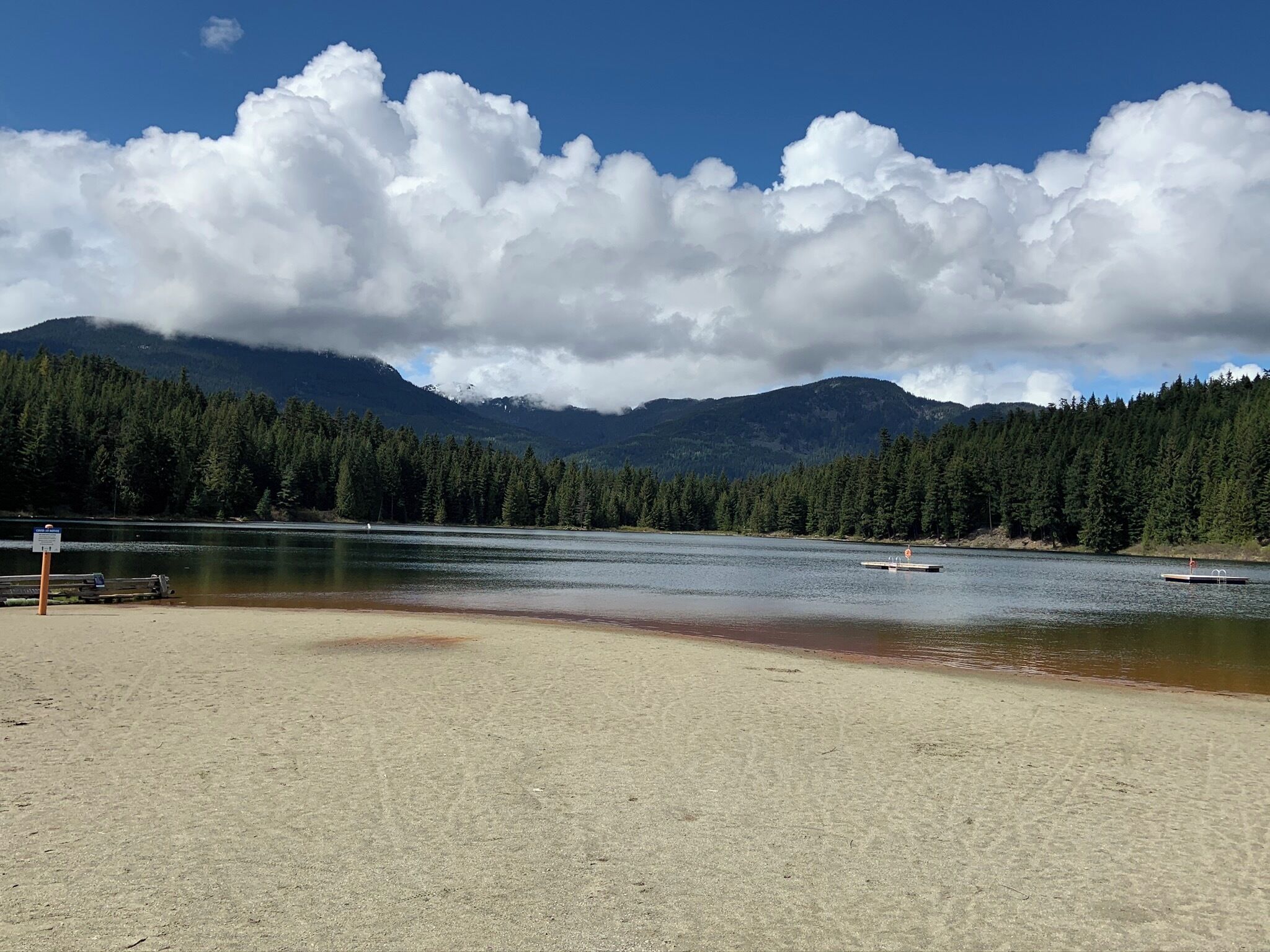 A beach with a wooden dock extending into a calm lake, surrounded by dense forest and mountains in the background.