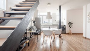 A modern apartment with a wooden staircase, a dining area with a round table and chairs, a kitchen with a washing machine, and a large window offering a city view. - Penthouse in Centro Historico with 2 Bedroom/2 Bathroom & Private Terrace (Centro)