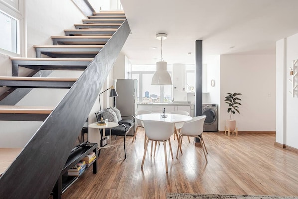 A modern apartment with a wooden staircase, a dining area with a round table and chairs, a kitchen with a washing machine, and a large window offering a city view.