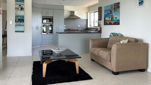 Une cuisine moderne avec des meubles gris, un four encastré et une fenêtre. Le salon est agrémenté d’un canapé beige et d’un tapis noir.