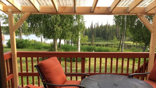 A covered patio with a table and chairs, overlooking a natural landscape with trees and a body of water. - Charlie’s house on the lake (Nikiski)