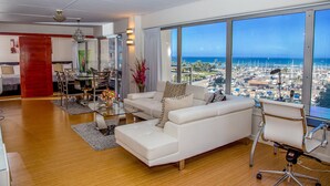A modern living room with a white sofa, glass coffee table, and a dining area with chairs and a table. A large window offers a view of a marina and cityscape. - Unobstructed Ocean View Suite. Don't miss out on the last-minute special rate! (Honolulu)