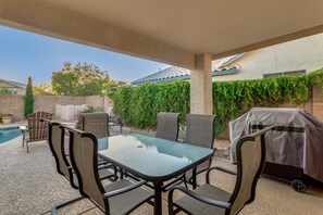 An outdoor patio with a glass-top table and chairs, a barbecue grill, and a swimming pool in the background.