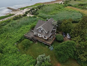 Une grande maison dotée d’une terrasse, entourée d’une végétation luxuriante et bordée par un plan d’eau.