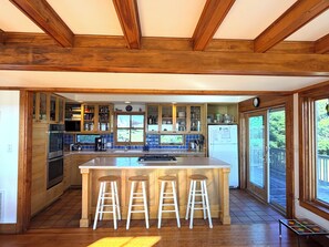 A kitchen with wooden beams, a large island with stools, and a view through the windows.
