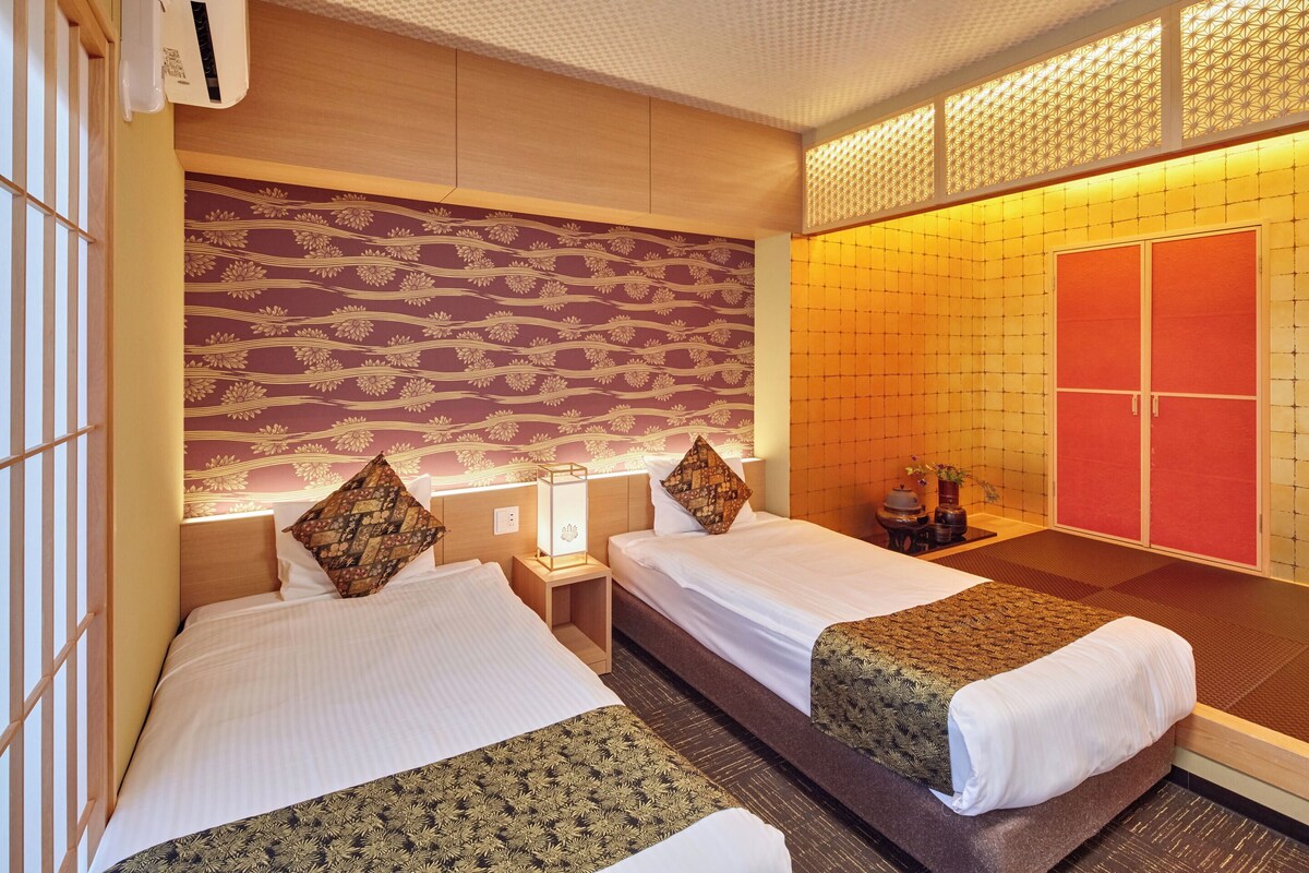 A hotel room with two beds, a nightstand, a wall-mounted lamp, a red wardrobe, and a patterned wall.