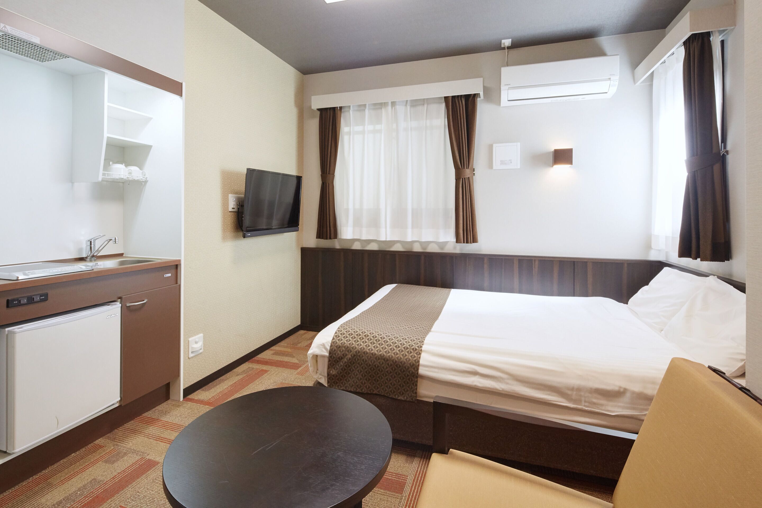 A hotel room with a bed, a desk, a chair, a small refrigerator, a TV mounted on the wall, and a window with curtains.