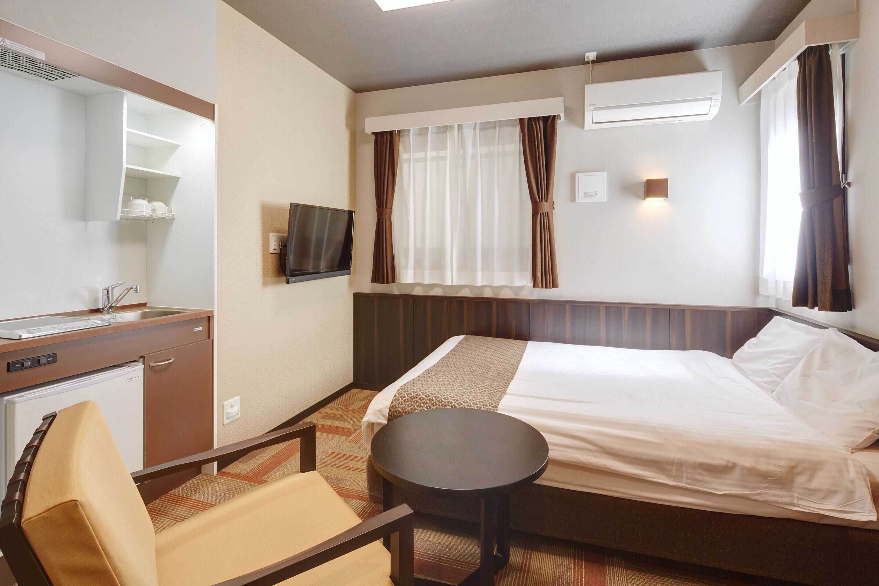 Habitación de hotel con cama, escritorio con refrigerador, silla, mesa redonda pequeña, televisor montado en la pared y ventana con cortinas.