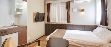 Habitación de hotel con cama, escritorio con refrigerador, silla, mesa redonda pequeña, televisor montado en la pared y ventana con cortinas.
