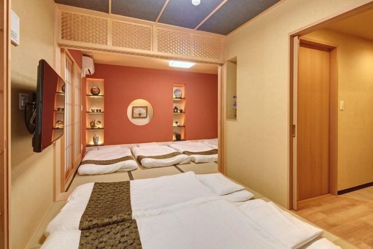 Ein Zimmer mit mehreren Betten, die in einer Reihe angeordnet sind, einem an der Wand befestigten Fernseher und Regalen mit verschiedenen Gegenständen.