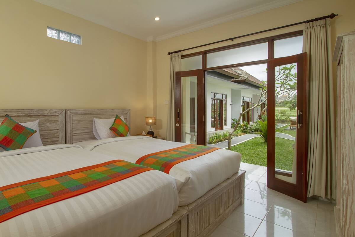 A bedroom with two beds, a wooden headboard, and a view of a garden through a sliding glass door.